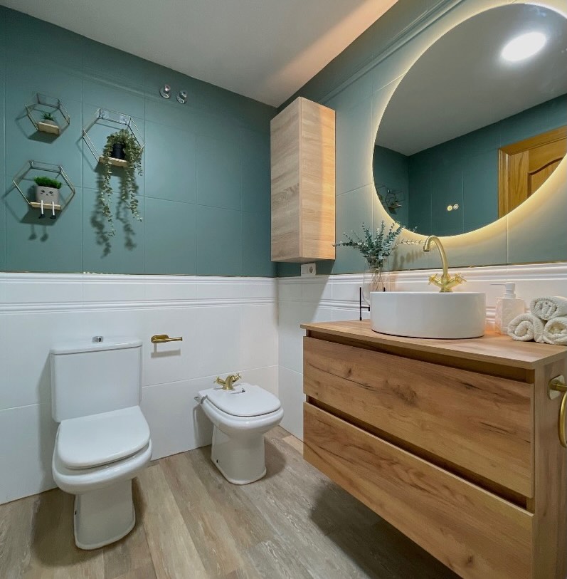 Baño reformado con pared verde, mueble de madera y espejo circular retroiluminado